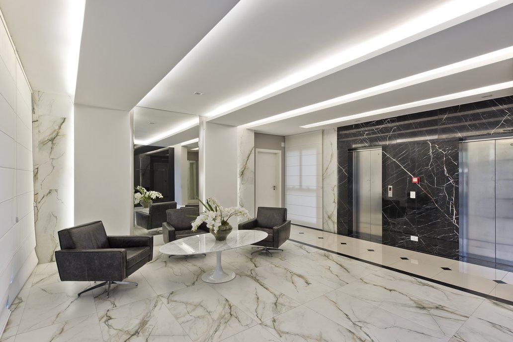 Com o conceito de continuidade, o porcelanato Bianco Paonazzetto prolonga do piso à parede e dá o maior acabamento do ambiente. O gesso com iluminação prolonga do teto à parede e o pórtico do elevador em preto facilita o entendimento do visitante quanto à localização no espaço.
