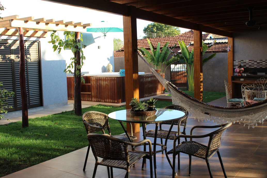 A estrutura de madeira é coberta com um forro de desenhos cortados a laser para criar um efeito de iluminação interessante. Além disso, esta cobertura protege do sol e da chuva, conformando um espaço perfeito para os mais variados climas. Faça chuva ou faça sol, os moradores desta casa podem curtir o dia ao ar livre! Seus pilares laterais são aproveitados para pendurar uma rede de balanço que além de dar um toque charmoso, ainda separa duas áreas de estar, criando uma ambientação.