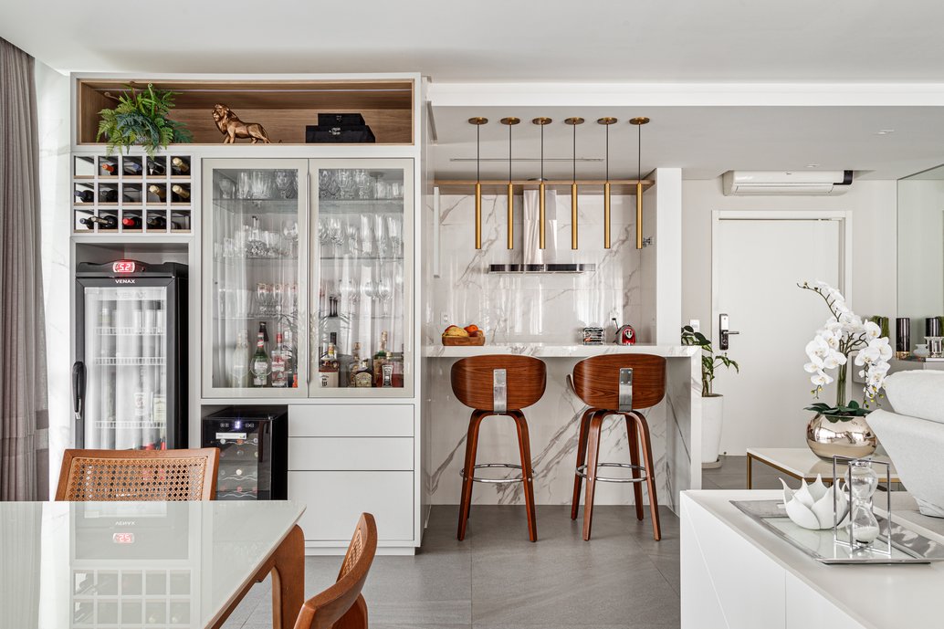 Em conjunto com o espaço para o bar, precisávamos pensar em um local para o casal acomodar sua cervejeira e adega. A solução neste caso foi a construção de um móvel funcional, bonito, com mix de texturas e materiais, para agregar os eletrodomésticos, sem perder a beleza. Inserimos portas em vidro na cristaleira para valorizar as taças, criamos um nicho superior amadeirado para decoração, e uma estrutura para comportar a coleção de bebidas.