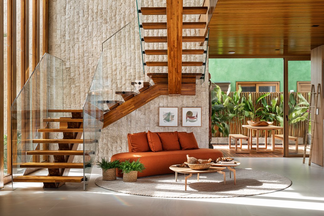 Escadaria com guarda-corpo em vidro, que emoldura o ambiente de espera, destacando-se através dos tons terrosos e texturas naturais. 
