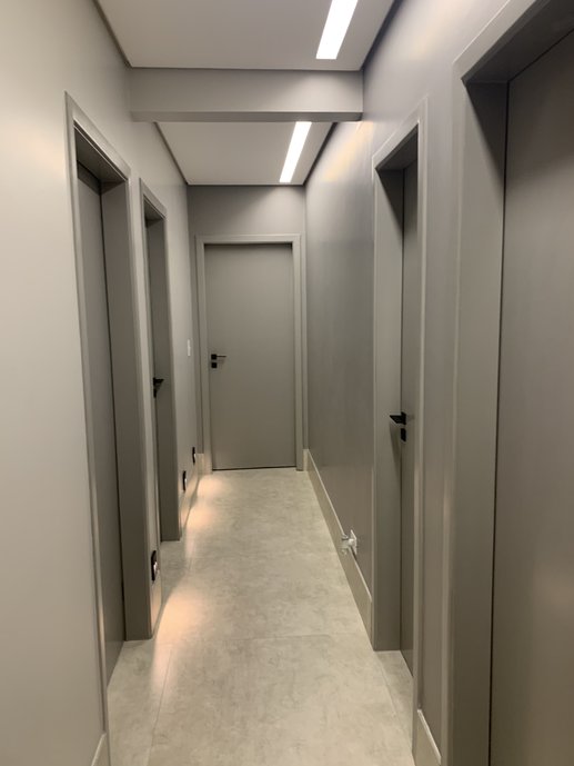 Corredor clean com paredes e portas cinzas, iluminação embutida no forro de gesso e balizaddores slim acima do rodapé. Porcelanato e rodapé da linha Broadway Lime da Portobello.