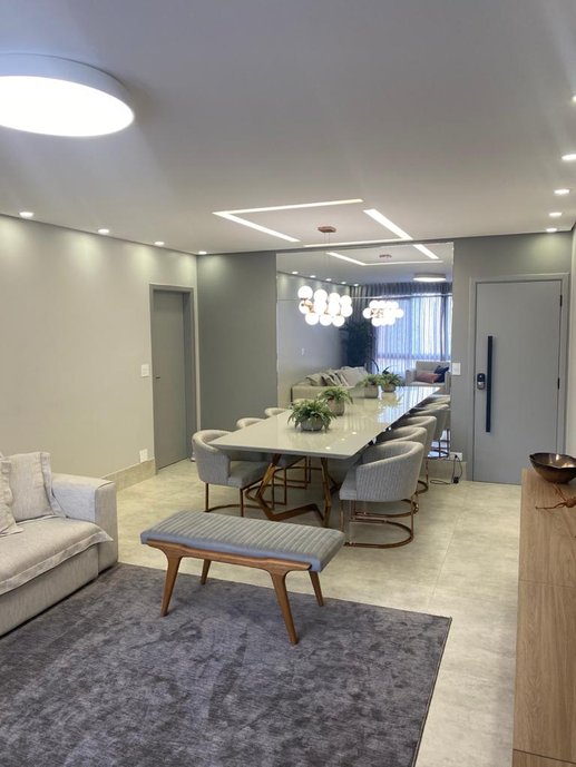 Salas de estar e jantar integradas, com paredes e portas cinzas, mesa e cadeiras com pés de inox rosê gold, e grande espelho para conferir maior amplitude. O tapete delimita o espaço do estar.