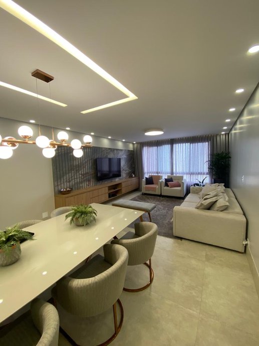 Salas de jantar e estar integradas, em tons de base neutras, como o cinza e o cru que, em contraste com o madeirado do home theater, transmitem aconchego e modernidade ao ambiente. Iluminação embutida em rasgos no forro de gesso e pendente acima da mesa de jantar, pequenas placas de led embutidas nas laterais e plafon na sala de estar. Todas as lâmpadas em tonalidades neutras que deixam o clima aconchegante e não modificam as cores do mobiliário.