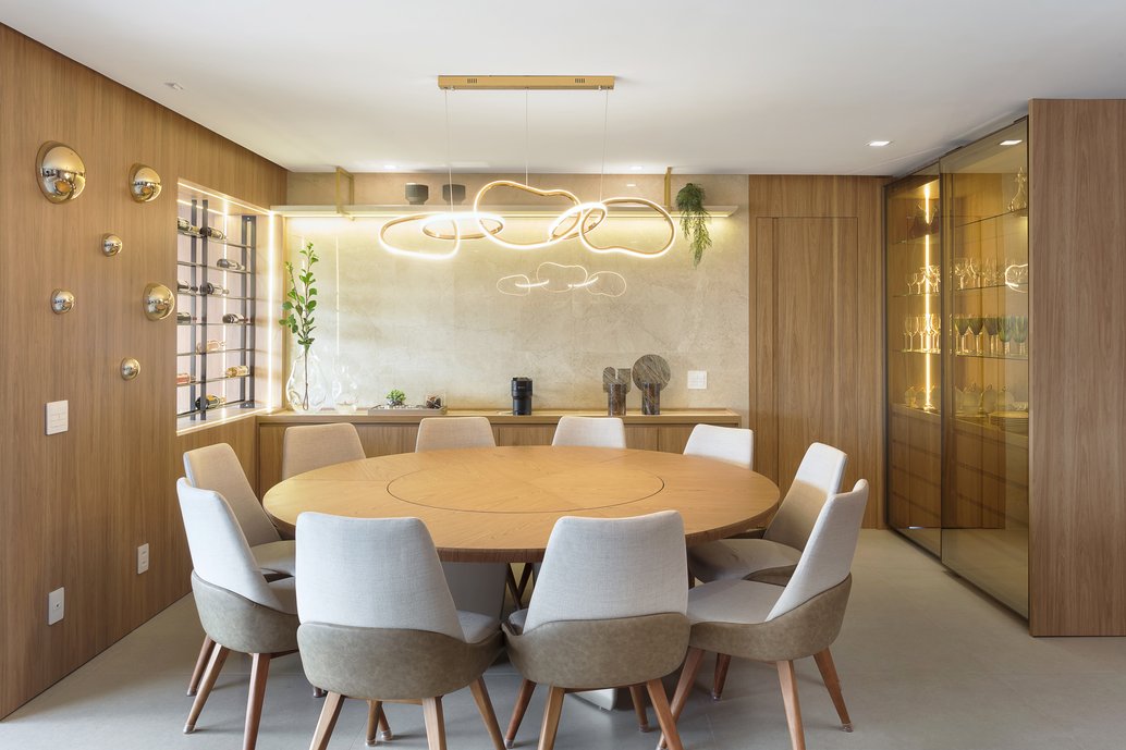 Sala de jantar moderna e sofisticada com mesa redonda de madeira, cadeiras estofadas, painel amadeirado, cristaleira iluminada, adega embutida e iluminação em LED. A combinação de madeira, pedra natural e tons neutros cria um ambiente aconchegante, elegante e atemporal. Inspire-se neste projeto de arquitetura de interiores para decorar salas de jantar contemporâneas, integradas e funcionais.