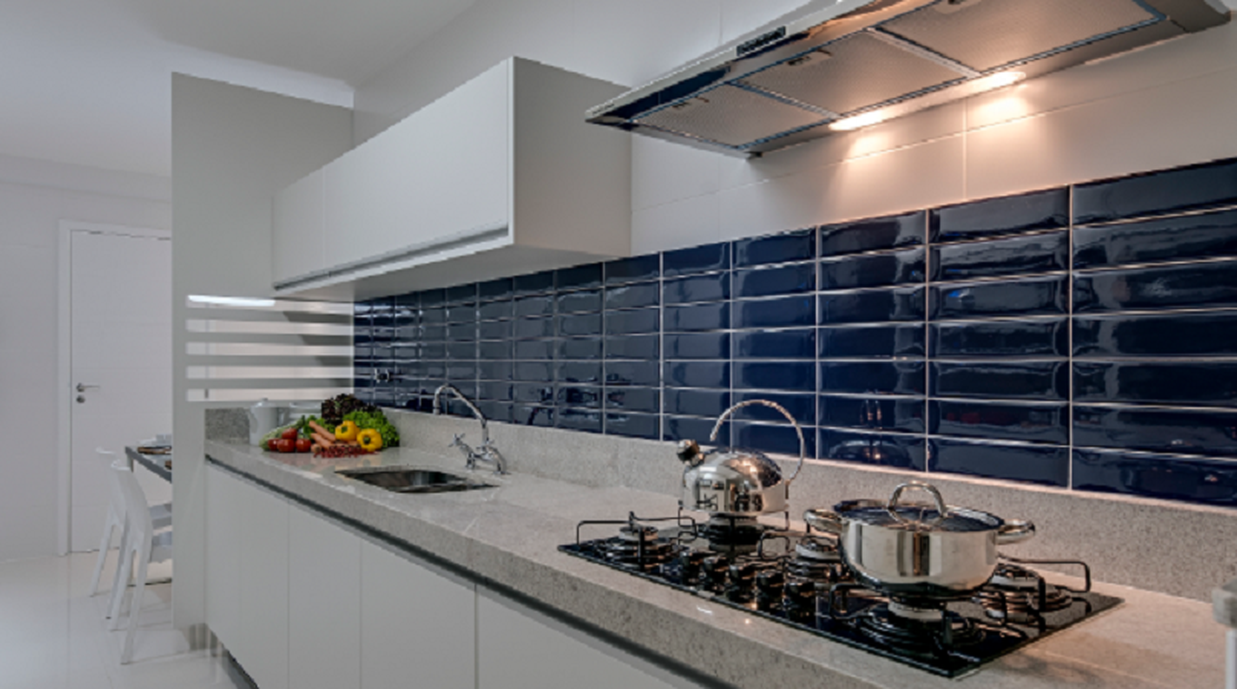 A faixa de revestimento de azulejos entre a bancada da pia e os armários suspensos, na linha retrô brilhante, deu o contraste perfeito para o branco puro do projeto. Portobello - Liverpool Azul Klein 7 X 24.4 cm.Esse formato retangular com bordas arrendondadas, nos remete aos azulejos dos metros ingleses criando um despojamento casual chique para o apartamento de praia de um executivo.